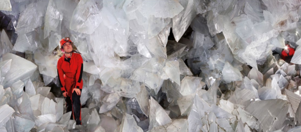 Giantgeodeofpulpí sians emporium Person in a red protective suit and helmet standing among jagged white crystals in a cavern